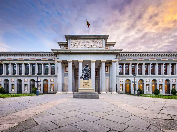 Prado Museum In Madrid Spain is a long white building with 6 large columns at the entrance, and a statue of a person sitting in a chair out front. 