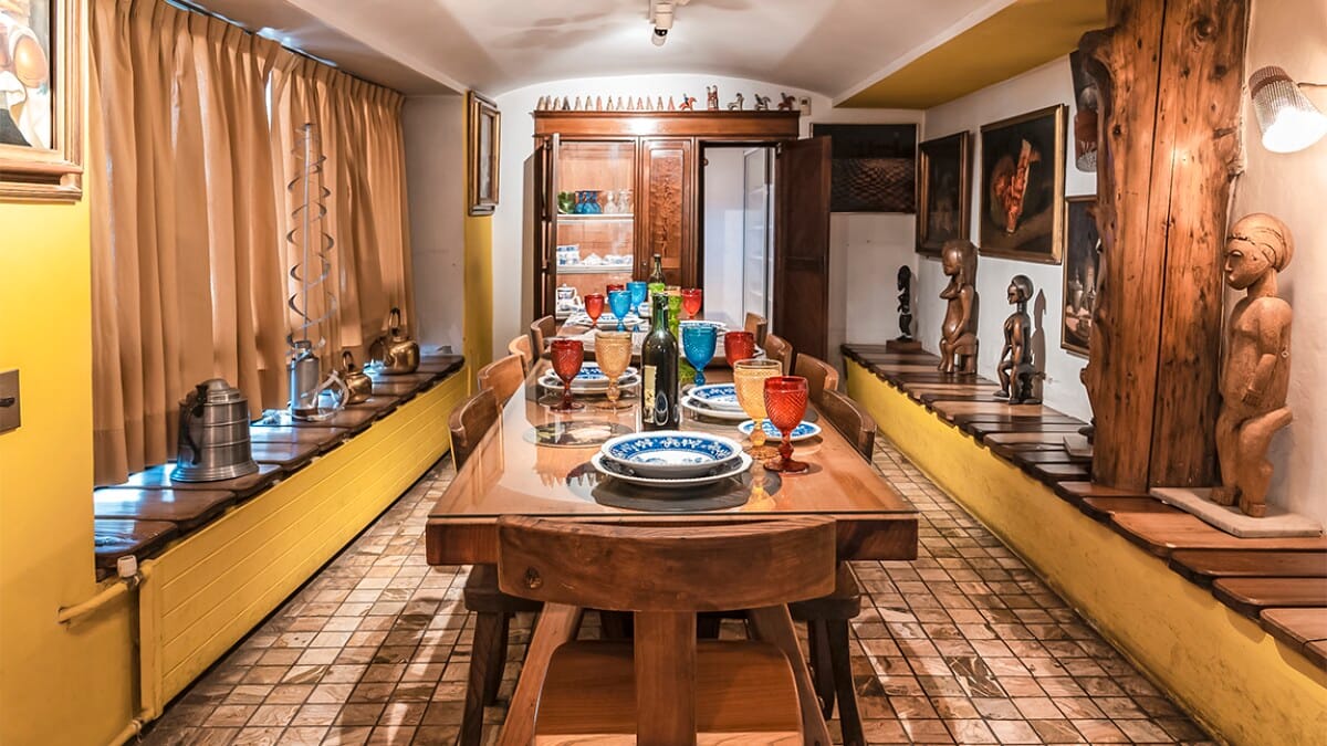 Dining room at La Chascona House Museum in Santiago, Chile, with a set table, colorful glassware, and wooden sculptures lining the walls.