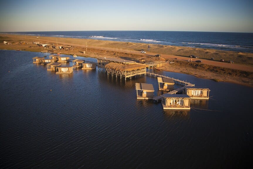 Aerial view of rustic wooden cabins built on stilts over the water at Laguna Garzón in Uruguay, with a sandy road, parked cars, and the Atlantic Ocean visible in the background during sunset.