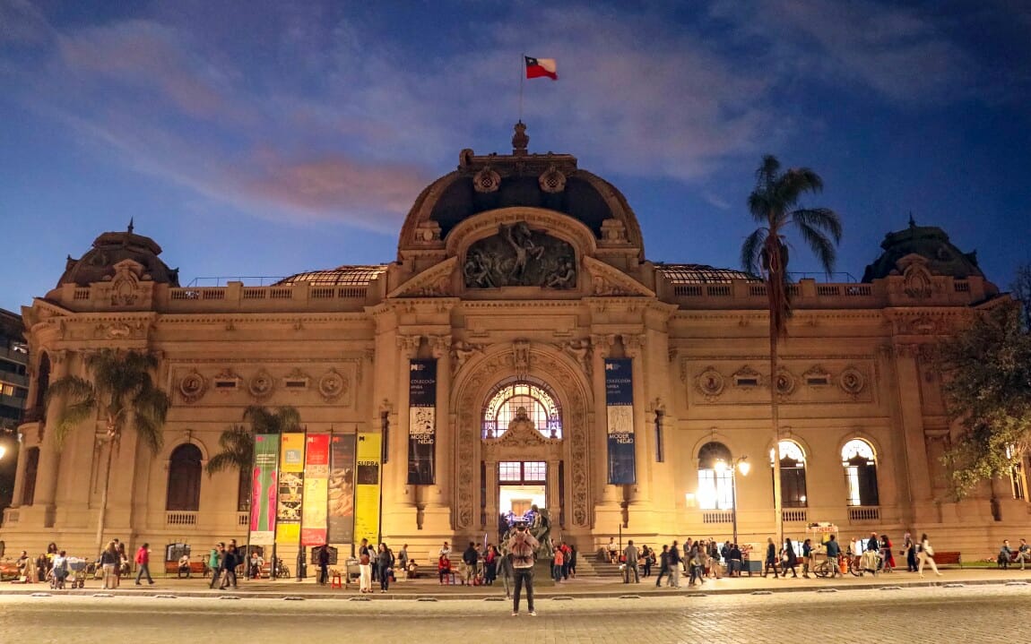 Night view of the National Museum of Fine Arts in Santiago, Chile, with people outside and the Chilean flag atop the ornate neoclassical building.