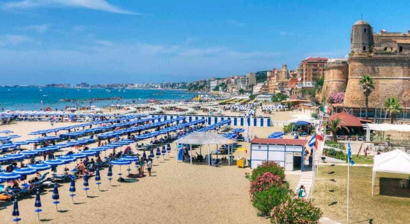 A beach near the edge of a city with people walking along the beach and there's lots of blue beach umbrellas along the beach.