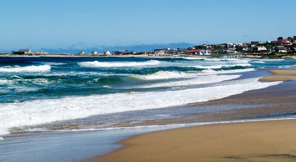 Waves rolling onto a sandy beach under a clear blue sky at Punta del Diablo, Uruguay, with a small coastal village featuring colorful houses and buildings in the background.