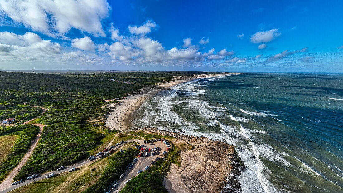 Aerial view of Santa Teresa National Park in Uruguay, featuring a wide sandy beach with ocean waves, a lush green forest, and a parking area near the shoreline under a bright blue sky with scattered clouds.