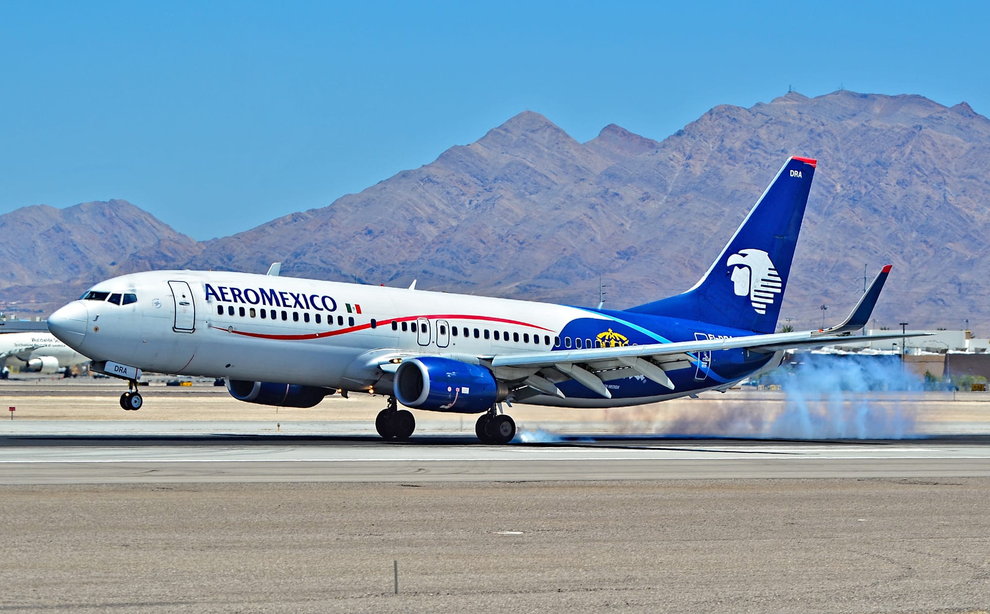Aeromexico aircraft touching down on a runway with smoke trailing from landing gear, mountains visible in the background.