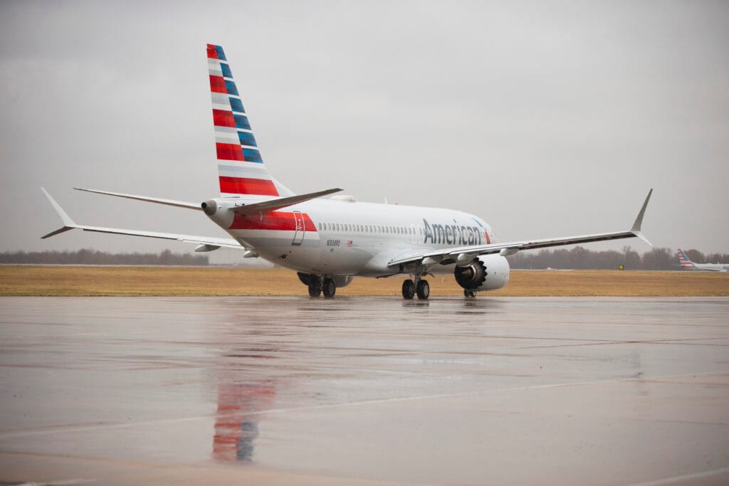 American Airlines Boeing 737 aircraft taxiing on a wet runway under overcast skies at an airport, with its tail fin displaying the red, white, and blue striped design.