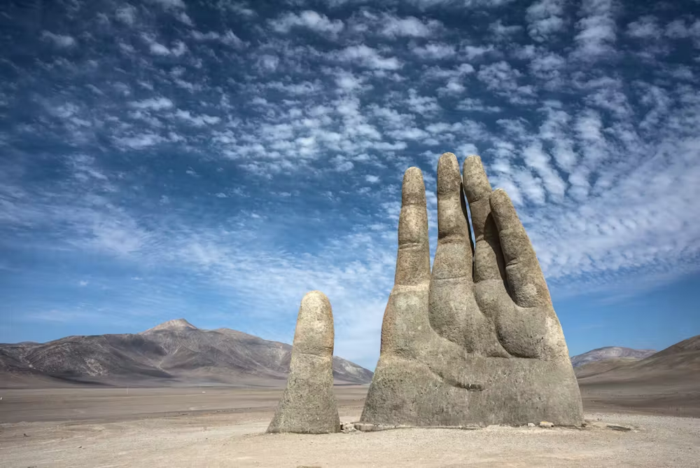 The Hand of the Desert sculpture rising from the sand in Chile’s Atacama Desert under a bright blue sky with scattered clouds.