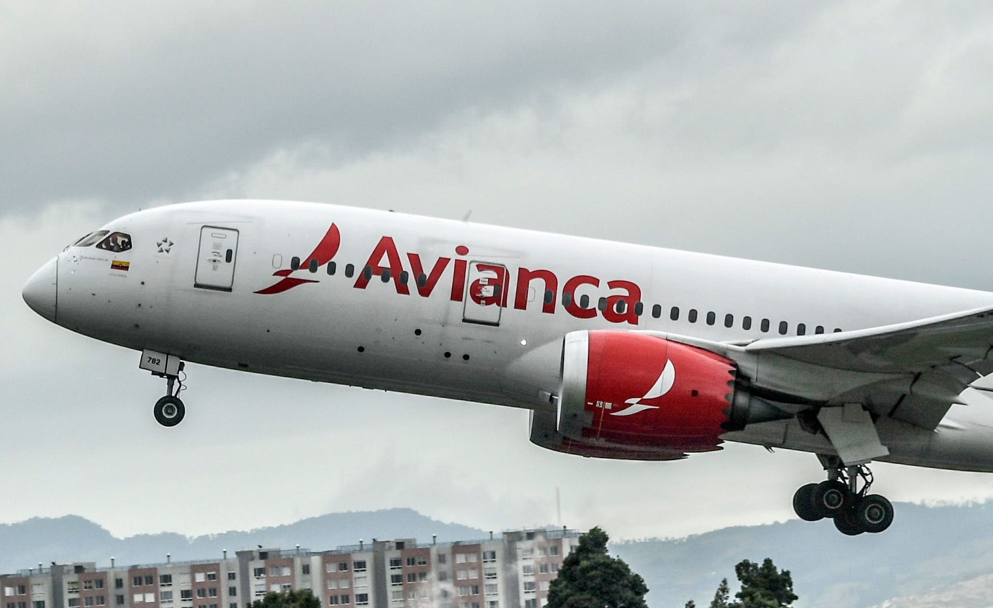 Avianca plane taking off and putting it's landing gear up, with mountains and an apartment complex seen in the background.