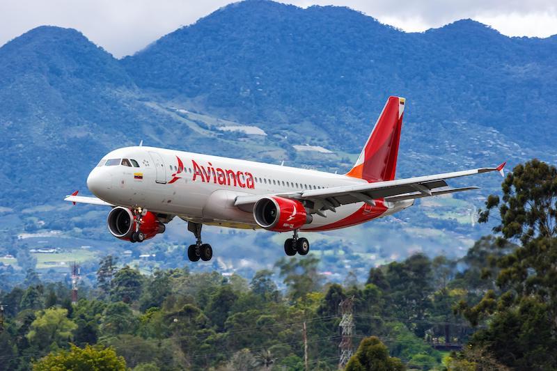 Avianca aircraft descending with landing gear deployed, flying over green terrain with mountains in the background.