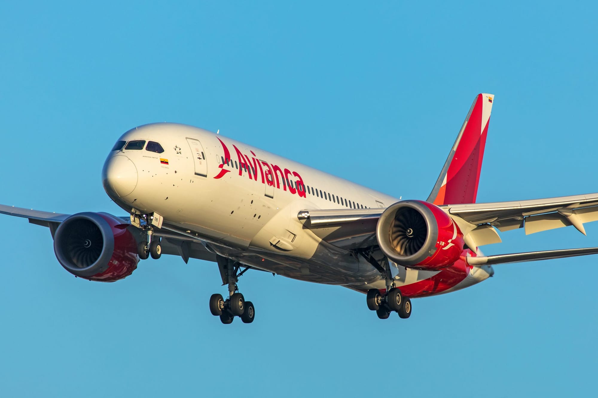 Avianca jetliner with red engine nacelles and tail fin preparing to land under clear blue sky