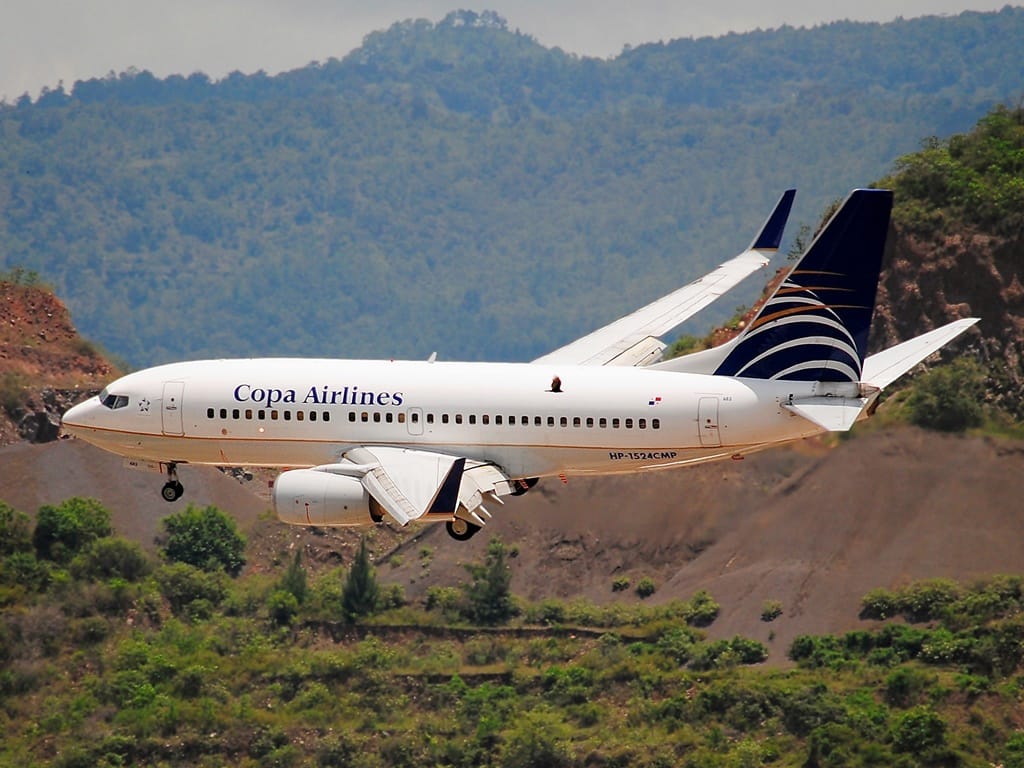 Copa Airlines aircraft on final approach for landing, flying over a hilly landscape with green vegetation.