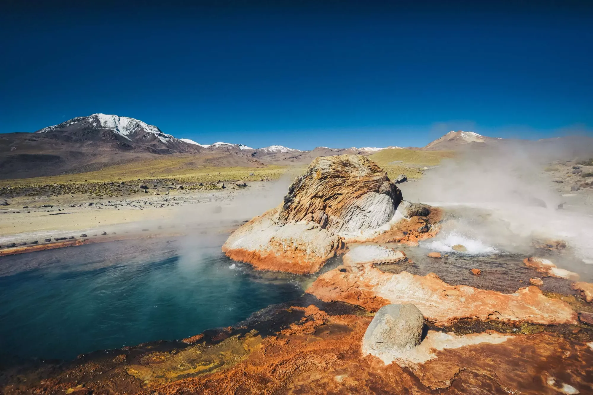 Steaming geothermal pool with vibrant orange mineral deposits and a large rock formation at El Tatio Geysers in Chile, set against a backdrop of snow-capped mountains and a clear blue sky.