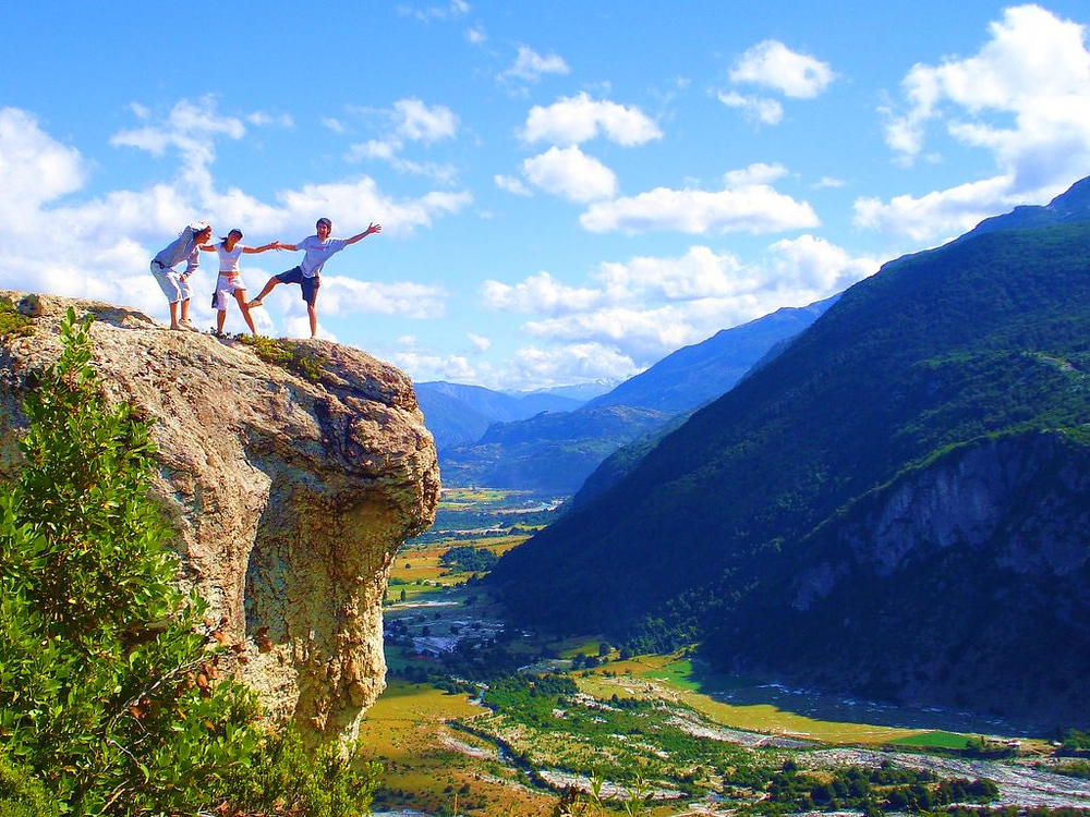 Three hikers posing playfully on the edge of Piedra del Águila, a large rocky outcrop overlooking a lush valley and surrounding green mountains in Chile under a bright blue sky with scattered clouds.