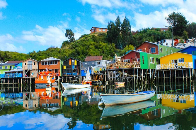 Colorful stilt houses and small boats reflected in calm water on Chiloé Island, Chile, with green hills and blue sky in the background.