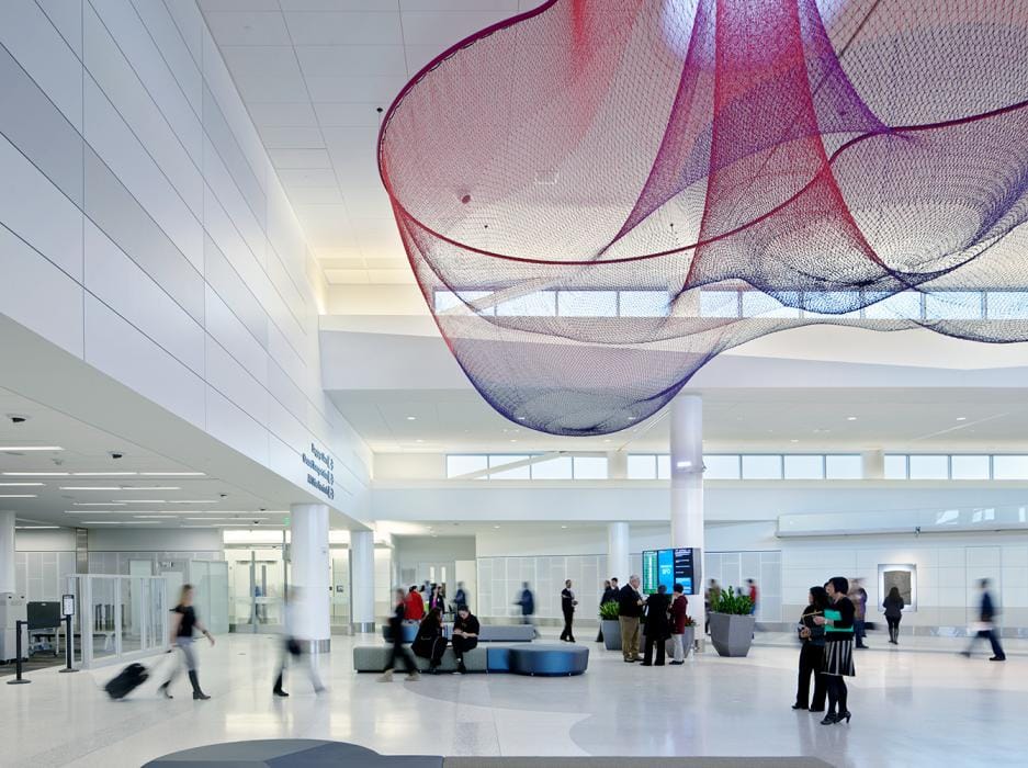 Travelers walking with luggage inside a modern white interior airport terminal with a red colored piece of art hanging from the ceiling at San Francisco International SFO Airport.