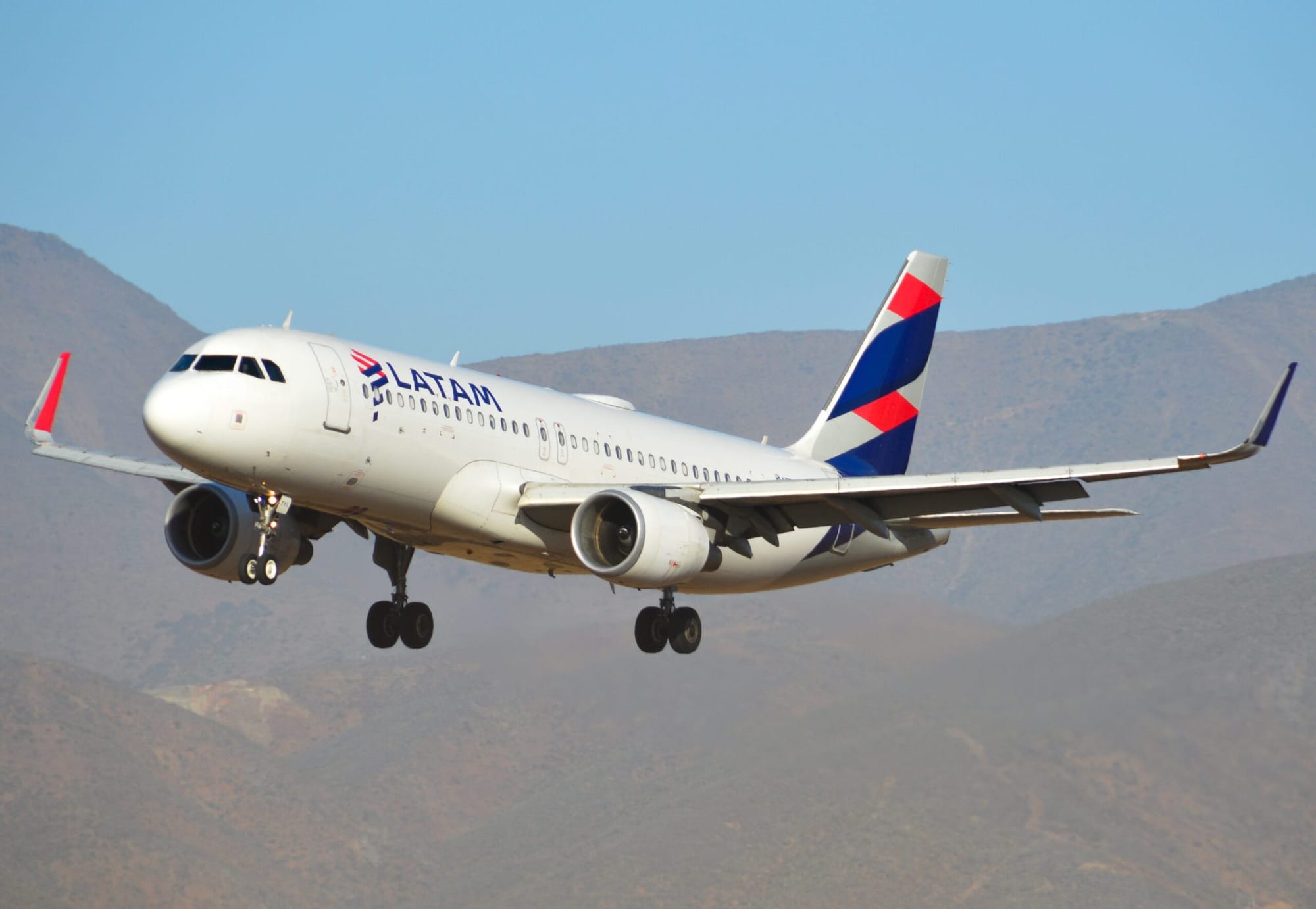 LATAM Airlines aircraft descending with landing gear extended, flying over mountainous terrain under a clear blue sky.