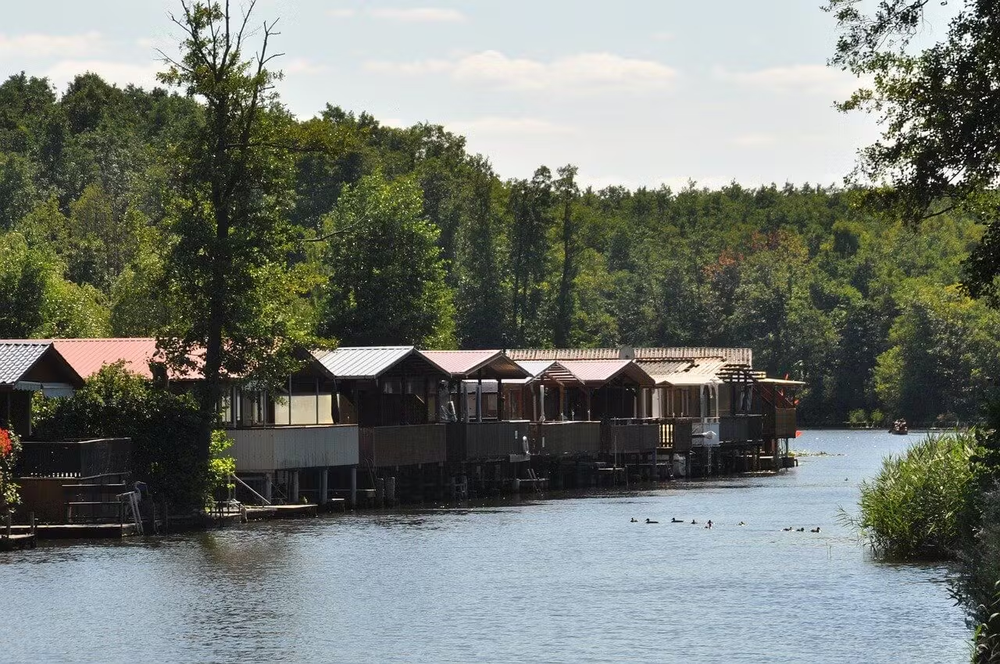Alt text: Row of lakeside floating cabins with sloped roofs along the edge of a calm waterway in Müritz National Park, surrounded by dense forest under a partly cloudy sky.