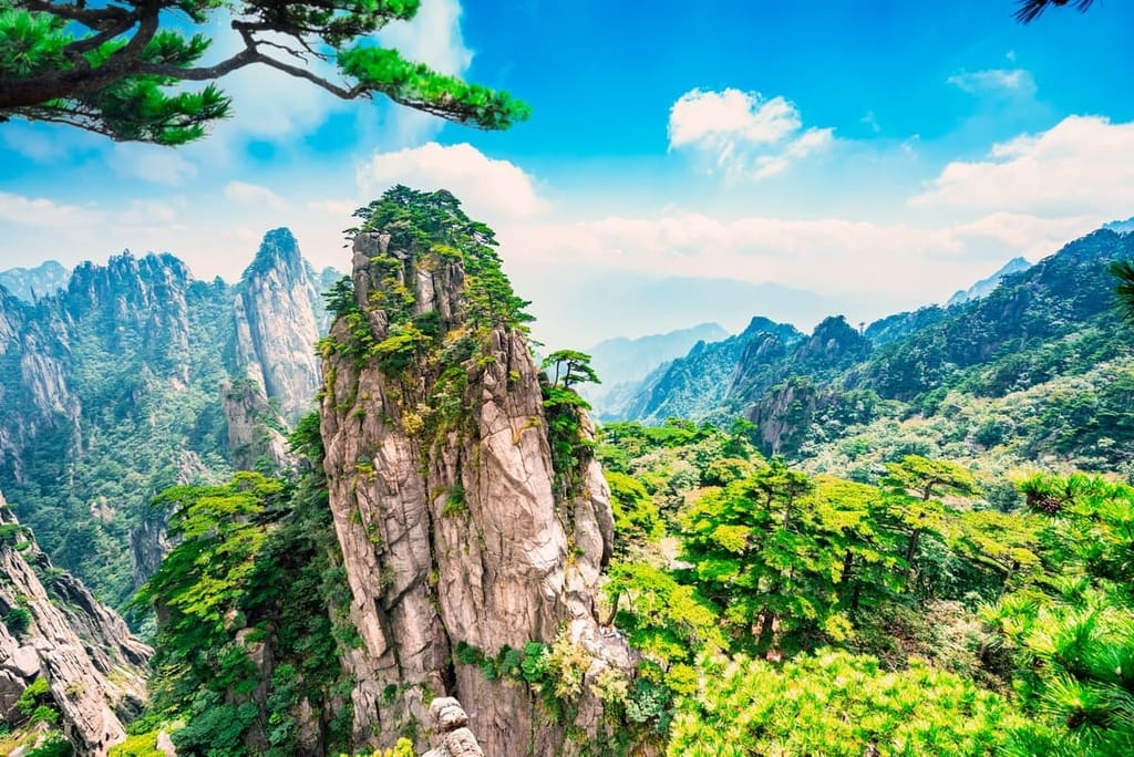 Bright daytime view of Mount Huangshan in Anhui, China, showcasing steep granite cliffs covered in green pine trees with a clear blue sky and distant mountain ranges in the background.
