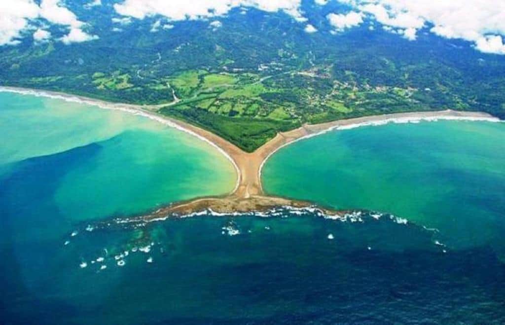 Aerial view of the famous Whale Tail sandbar formation at Playa Ballena in Marino Ballena National Park, Costa Rica, showing two curved beaches meeting at a central point surrounded by turquoise ocean waters and lush green coastline under a partly cloudy sky.