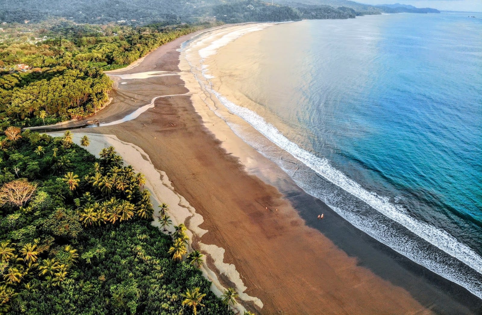 Aerial view of Playa Dominical near Uvita, Costa Rica, showing a long stretch of sandy beach lined with lush tropical vegetation, gentle ocean waves rolling onto the shore, and a backdrop of green hills under a soft, partly cloudy sky.