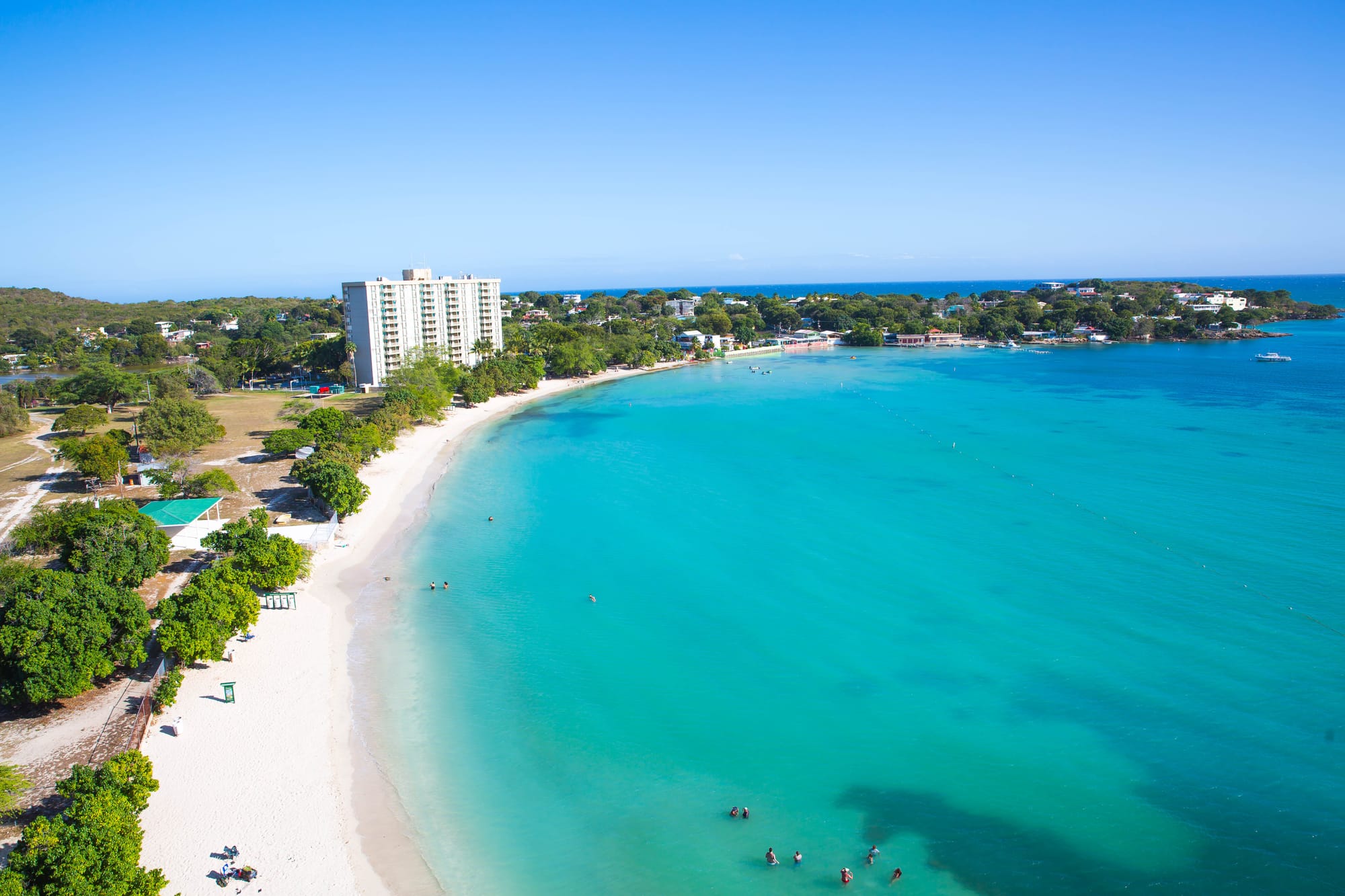 Aerial view of Playa Santa Beach near Ponce, Puerto Rico, featuring a curved white sandy shoreline, clear turquoise waters with swimmers, lush green trees, and a tall beachfront building under a bright blue sky.