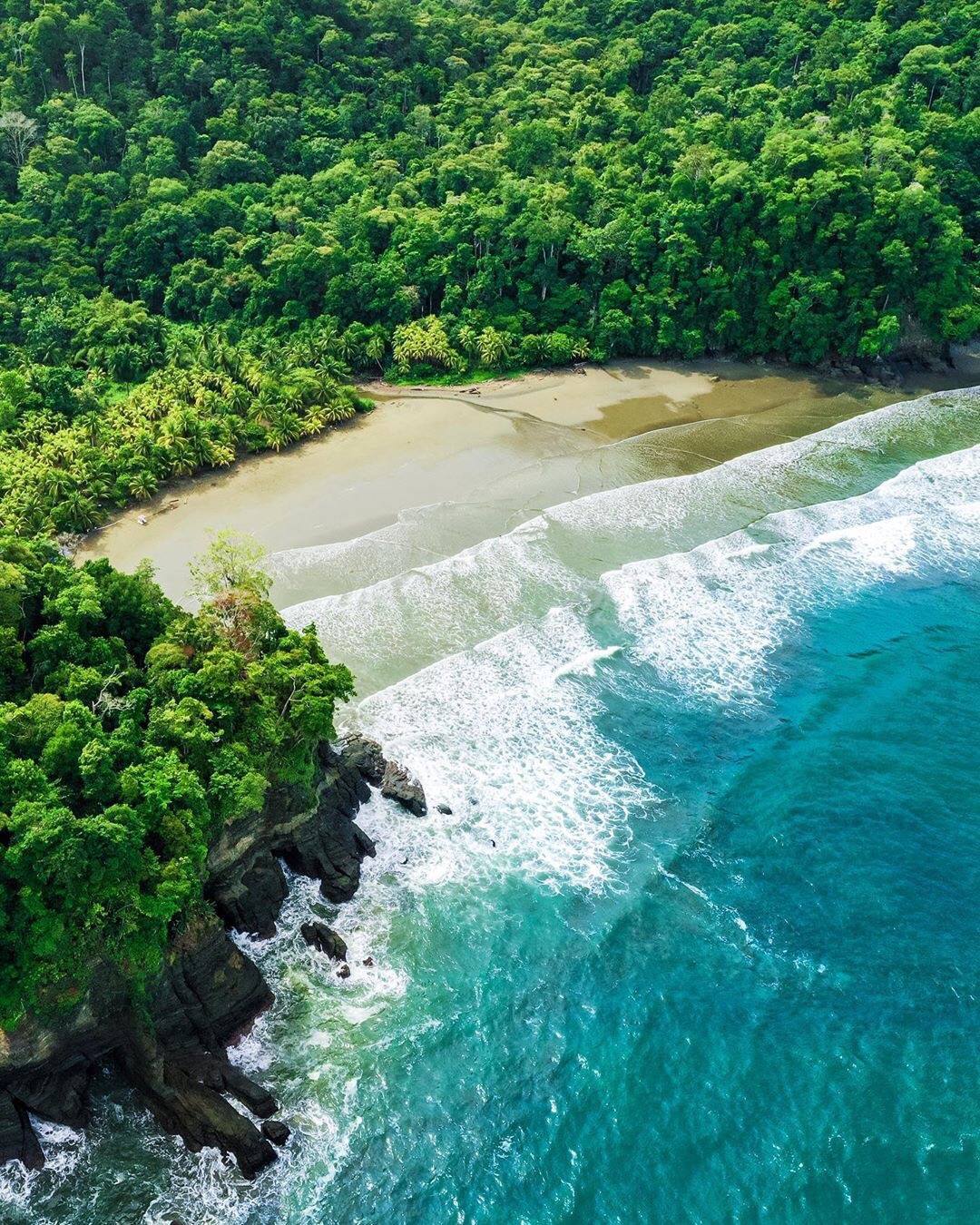 Aerial view of Playa Ventanas in Costa Rica, featuring a secluded sandy beach surrounded by dense tropical rainforest, rocky cliffs along the shoreline, and turquoise ocean waves crashing against the coast.