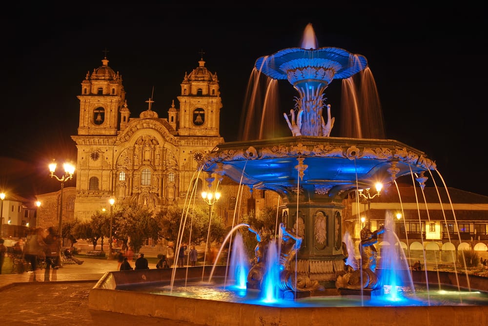 Illuminated colonial-era fountain with blue-lit water in Plaza de Armas, Cusco, Peru, with the historic Church of the Society of Jesus brightly lit in the background against the night sky.