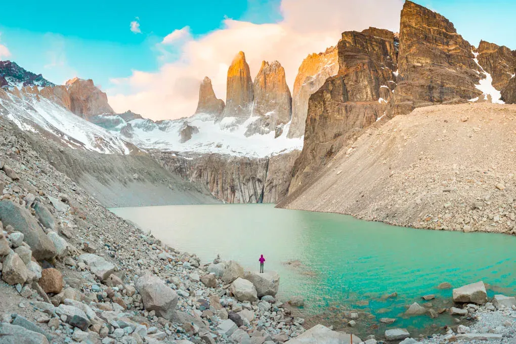Person standing by turquoise lake with rocky mountains and snowy peaks at Torres del Paine under a bright blue sky.