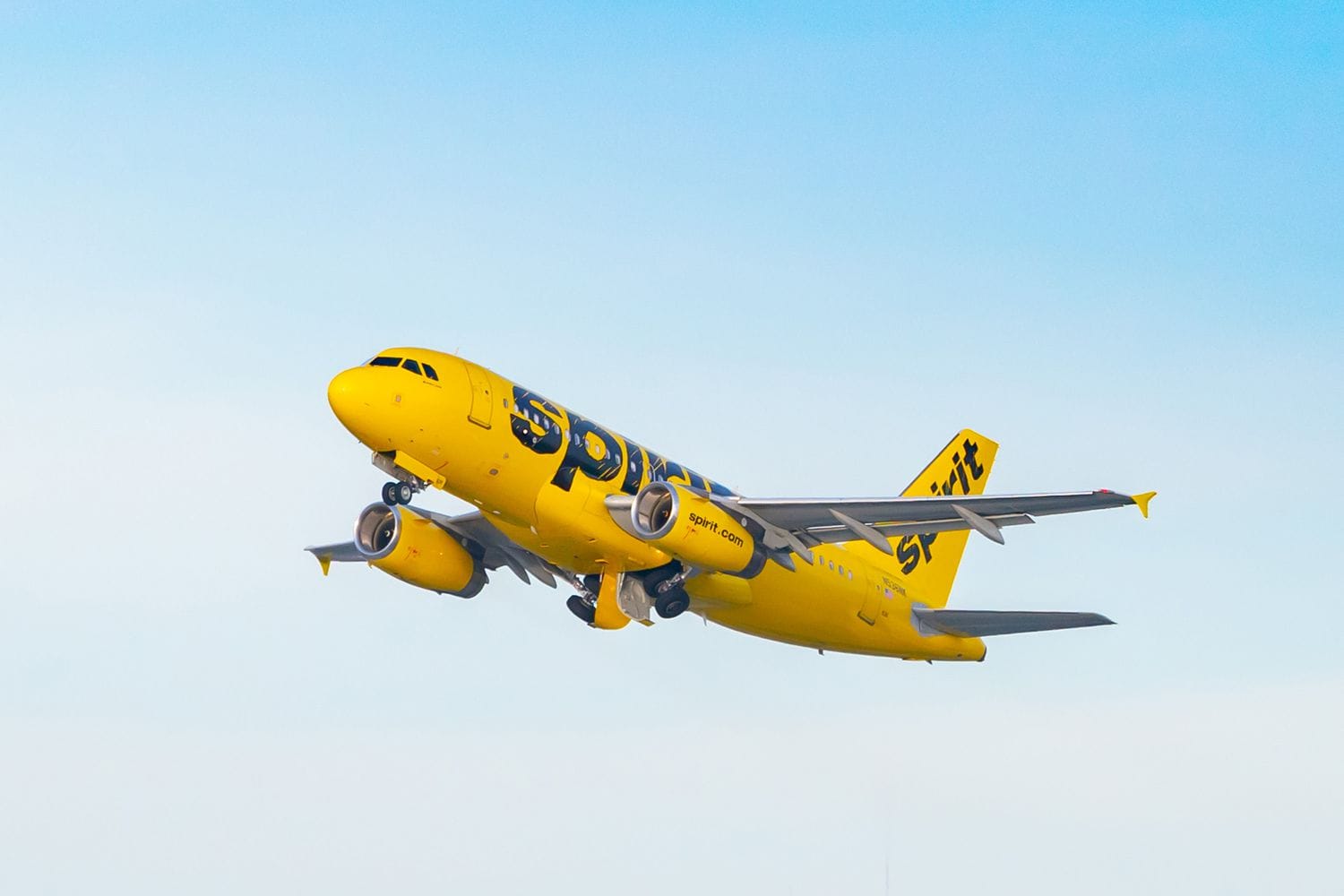 Bright yellow Spirit Airlines airplane ascending against a clear blue sky during takeoff or landing approach.