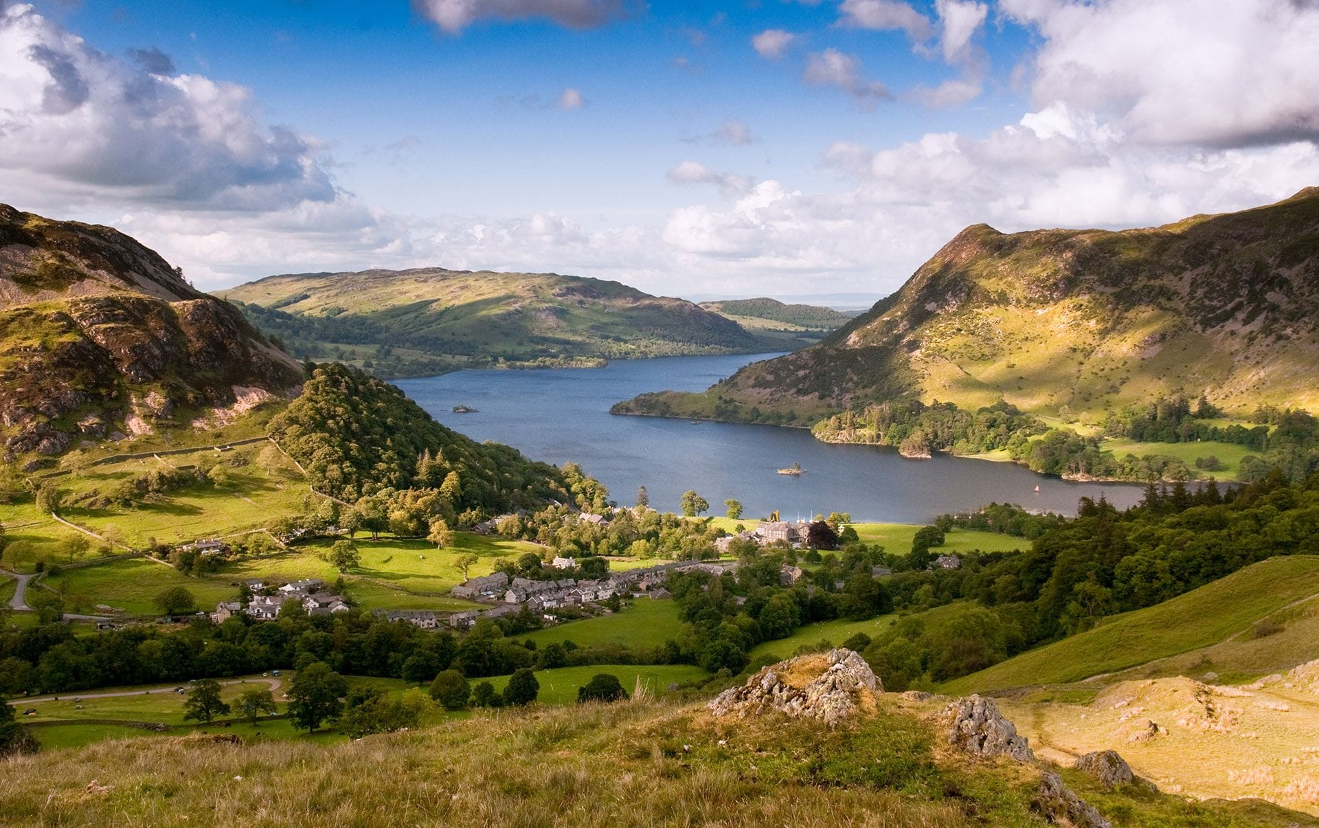 A scenic view of a village by a blue lake surrounded by green hills and rocky mountains under a partly cloudy sky.