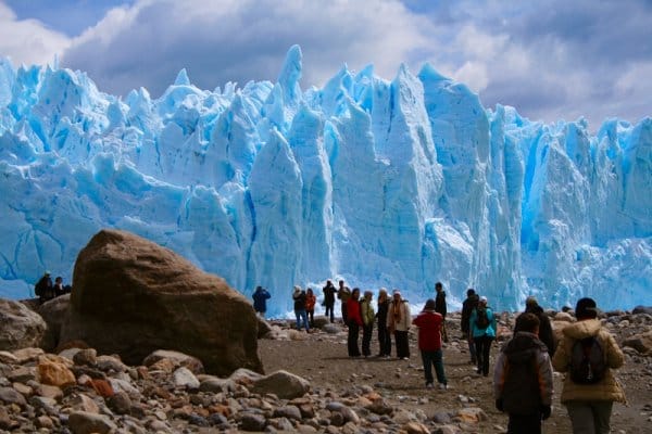 Tourists standing near the bright blue ice wall of a glacier in Torres del Paine National Park, Chile, with rocky ground and cloudy skies.