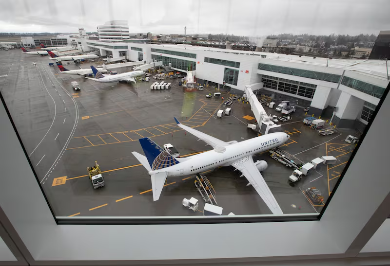 Aerial view of a United Airlines plane parked at a gate at Seattle-Tacoma International Airport (SEA) on a cloudy day, with multiple aircraft from various airlines, ground service vehicles, and terminal buildings visible across the tarmac.