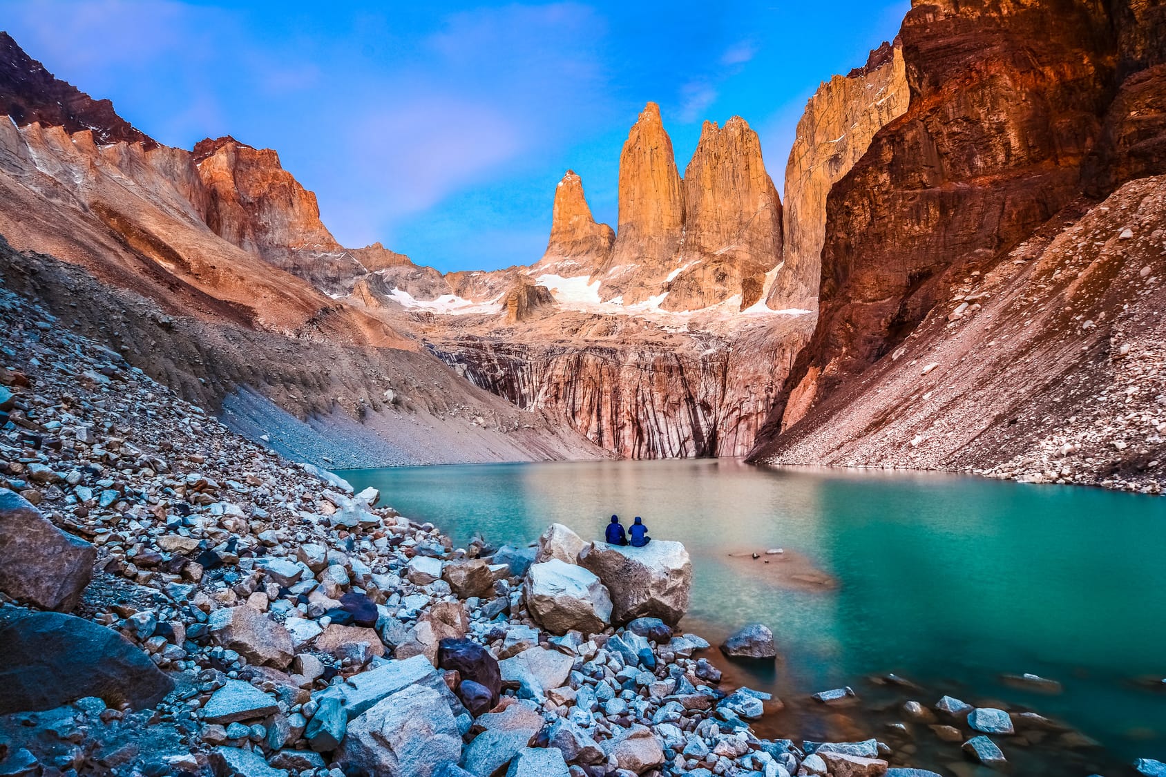 Two hikers sitting on rocks by a turquoise glacial lake at the base of the iconic Torres del Paine granite peaks in Torres del Paine National Park, Chile, under a vibrant blue sky.