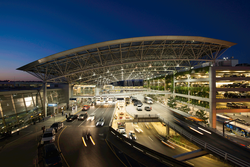Exterior view of the entrance to Portland International Airport (PDX) at dusk, showing a modern, well-lit terminal with a large curved steel and glass canopy.