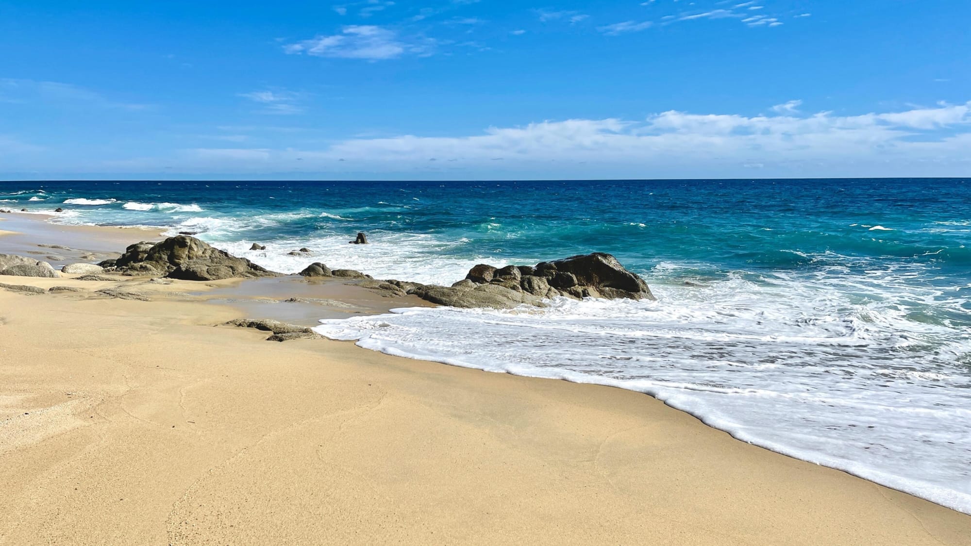 View of Playa Tortuga near Uvita, Costa Rica, featuring a sandy beach with scattered rocks along the shoreline, turquoise waves crashing gently, and a bright blue sky with light clouds overhead.