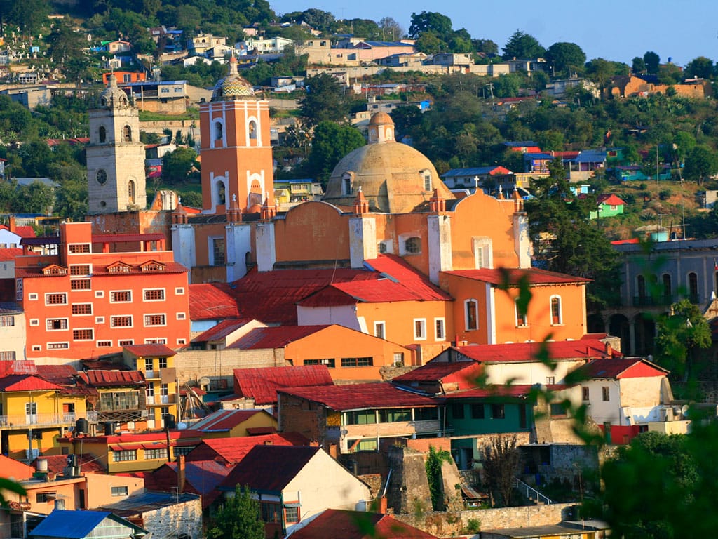 Colorful colonial buildings with red rooftops and a large domed church in the historic town of Batopilas, Mexico, surrounded by lush green hills and scattered homes.