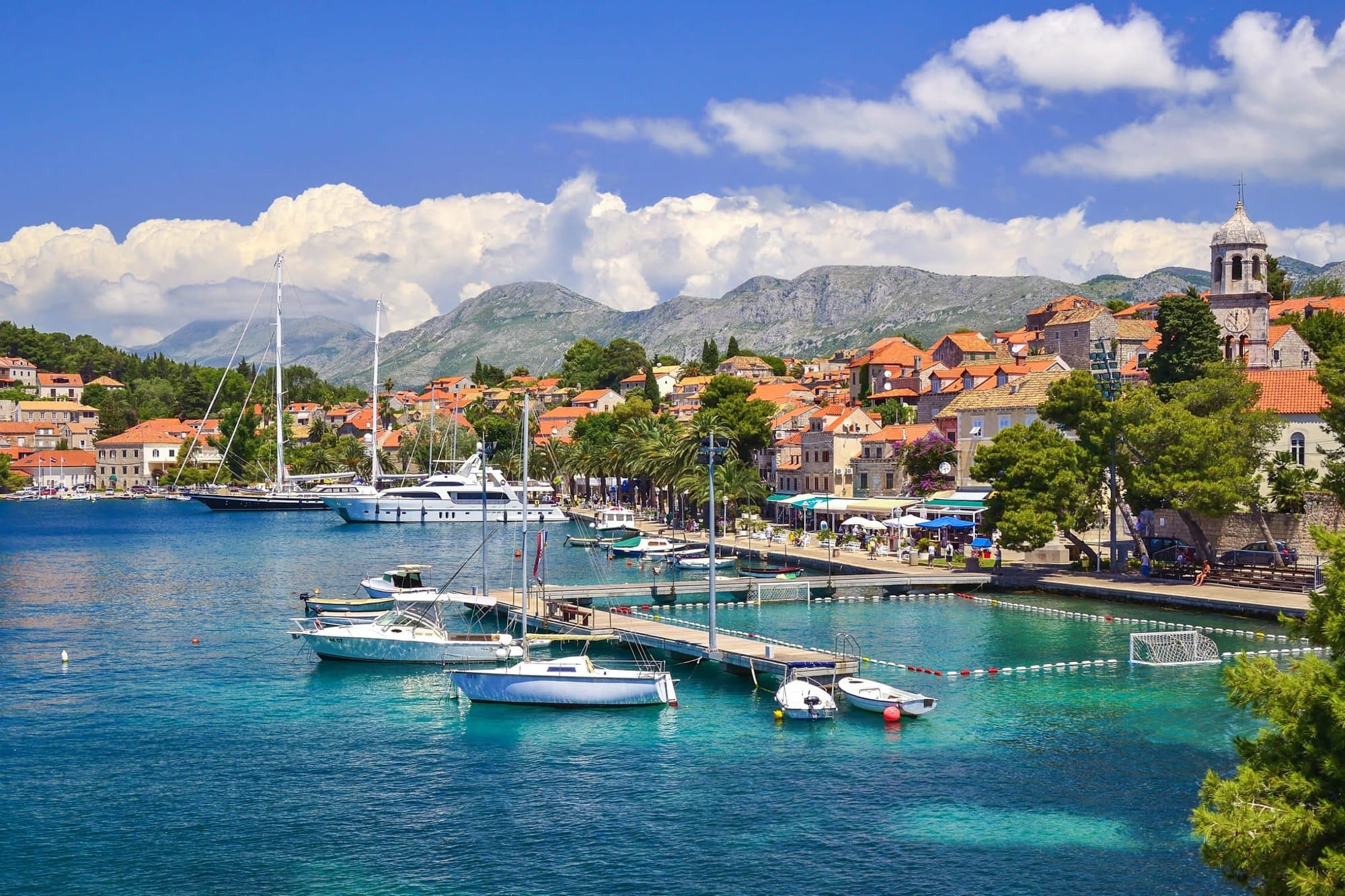 Scenic waterfront of Cavtat, Croatia, with yachts and boats docked along the marina, palm-lined promenade, and terracotta-roofed buildings nestled against mountainous terrain.