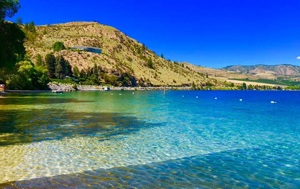 Calm, transparent waters of Lake Chelan against rolling hills and a cloudless sky.