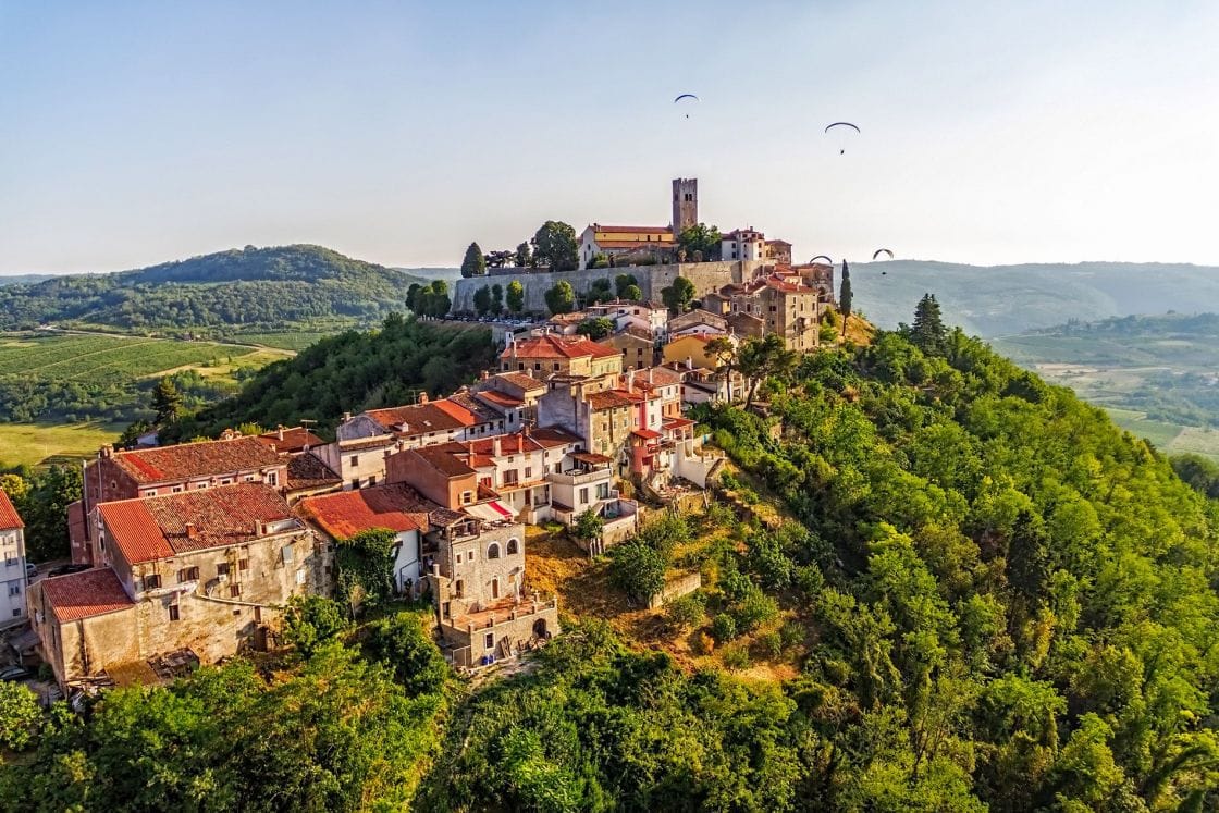 Picturesque medieval street in Grožnjan, Croatia, with old stone buildings, flowerpots, hanging plants, bicycles, and rustic wooden barrels lining a cobbled path beneath a sunny sky.
