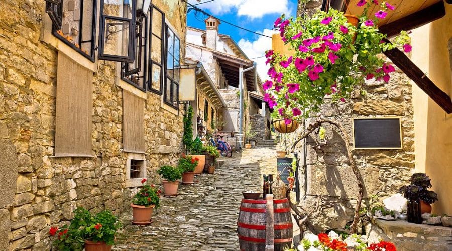 Narrow cobblestone alleyway in Hum, Croatia, the world’s smallest town, lined with rustic stone buildings, flower pots, open windows, and colorful hanging flowers under a bright blue sky.