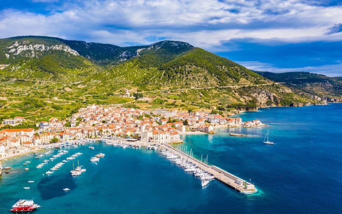 Aerial view of Komiža, Croatia, a seaside village on Vis Island with orange-roofed stone houses, vibrant blue water, a marina filled with boats, and green hills in the background.