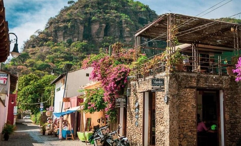 Colorful cobblestone street in Malinalco, Mexico, lined with shops, flowering bougainvillea, and a rustic stone building with a rooftop terrace, set against a backdrop of lush green hills.
