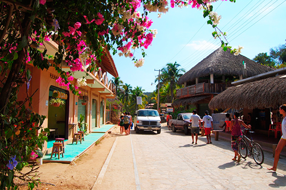 Vibrant street scene in Mazunte, Mexico, with people walking and biking past pastel-colored buildings and thatched-roof restaurants, framed by blooming bougainvillea and palm trees.