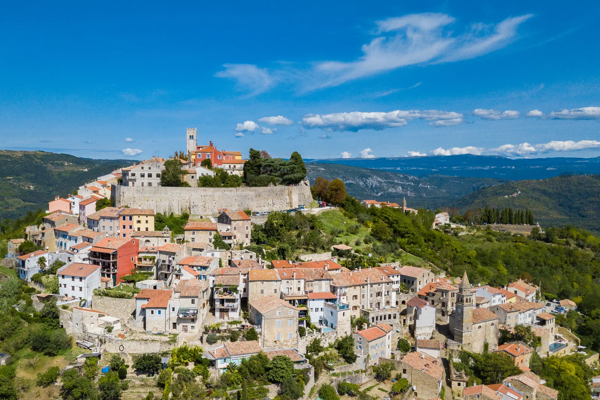 Aerial view of the hilltop town of Motovun, Croatia, featuring medieval stone buildings with red-tiled roofs clustered within ancient city walls, surrounded by green forested hills and a bright blue sky.