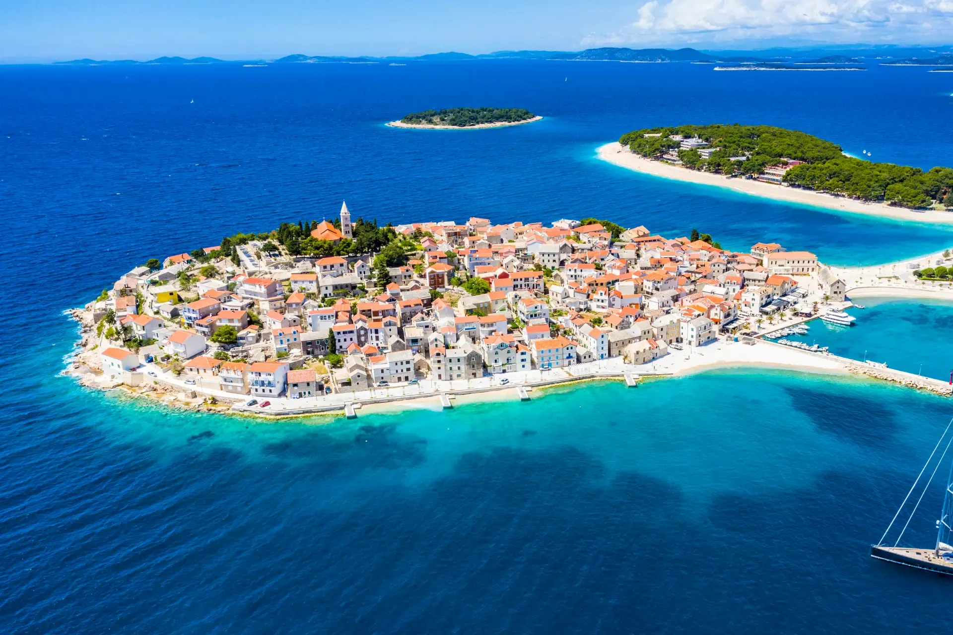 Aerial view of the coastal town of Primošten, Croatia, located on a small peninsula surrounded by turquoise waters, with tightly packed red-roofed buildings and nearby green islands.