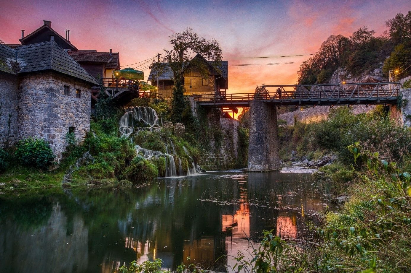 Serene riverside view of Rastoke, Croatia, at dusk, with traditional stone houses, wooden bridges, and cascading waterfalls reflecting in the calm waters beneath a colorful sunset sky.