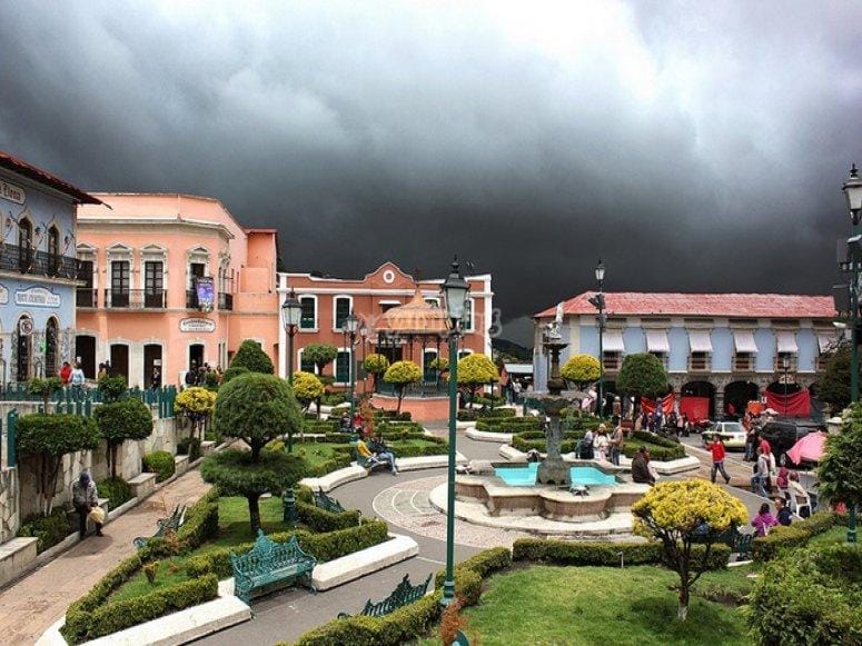 Picturesque town square in Real de Monte, Mexico, with manicured gardens, a central fountain, pastel-colored colonial buildings, and people strolling under a dramatic stormy sky.