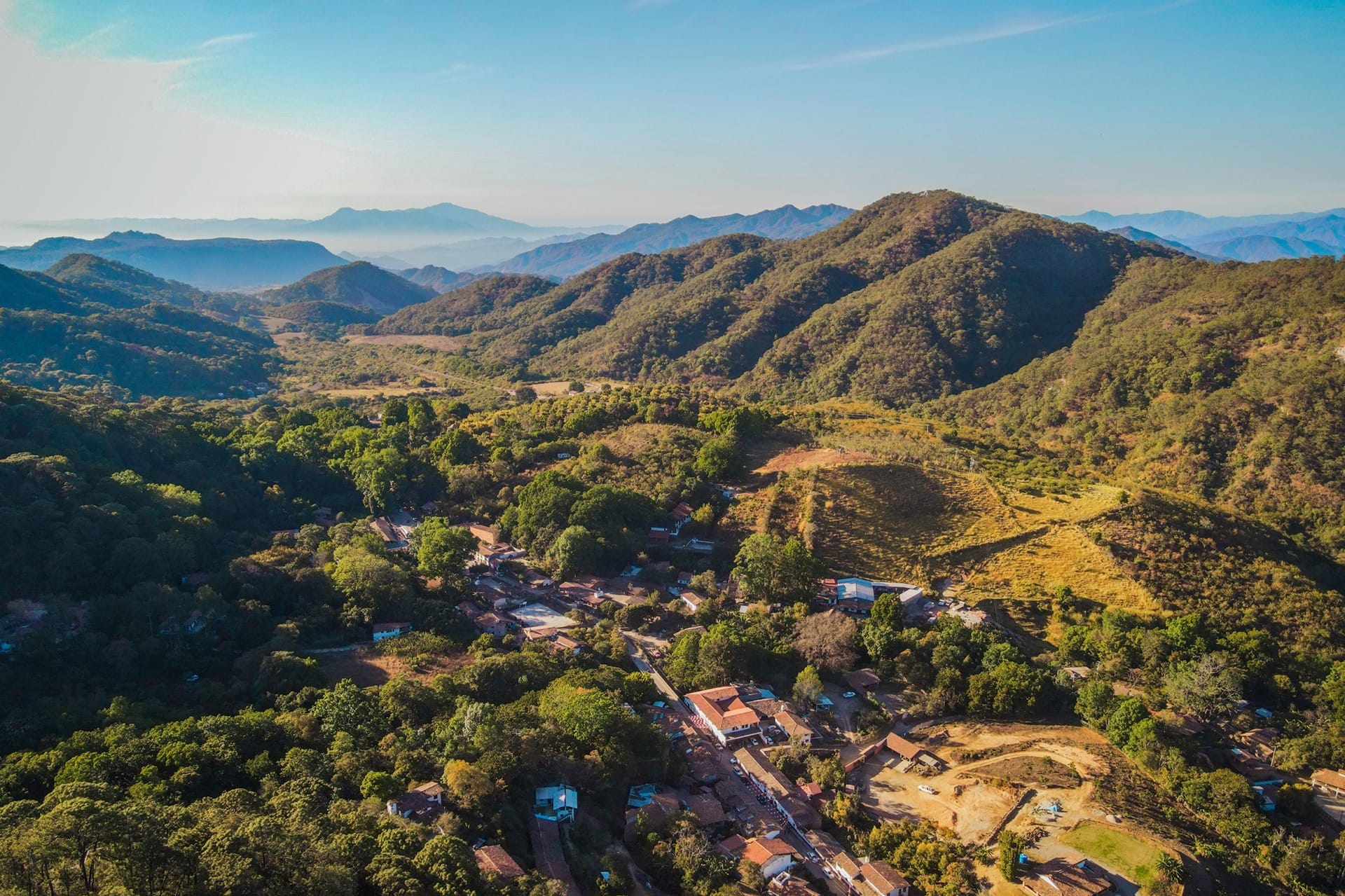 Aerial view of San Sebastián del Oeste, Mexico, a mountain village surrounded by dense forest and rolling hills, with a mix of tiled rooftops and rural farmland.