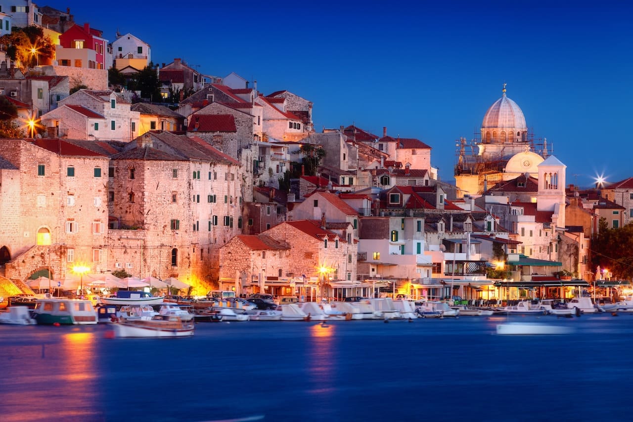 Evening view of Šibenik Old Town, Croatia, with historic stone buildings and red-tiled roofs stacked along the waterfront, illuminated by warm lights, with the Cathedral of St. James prominently in the background.