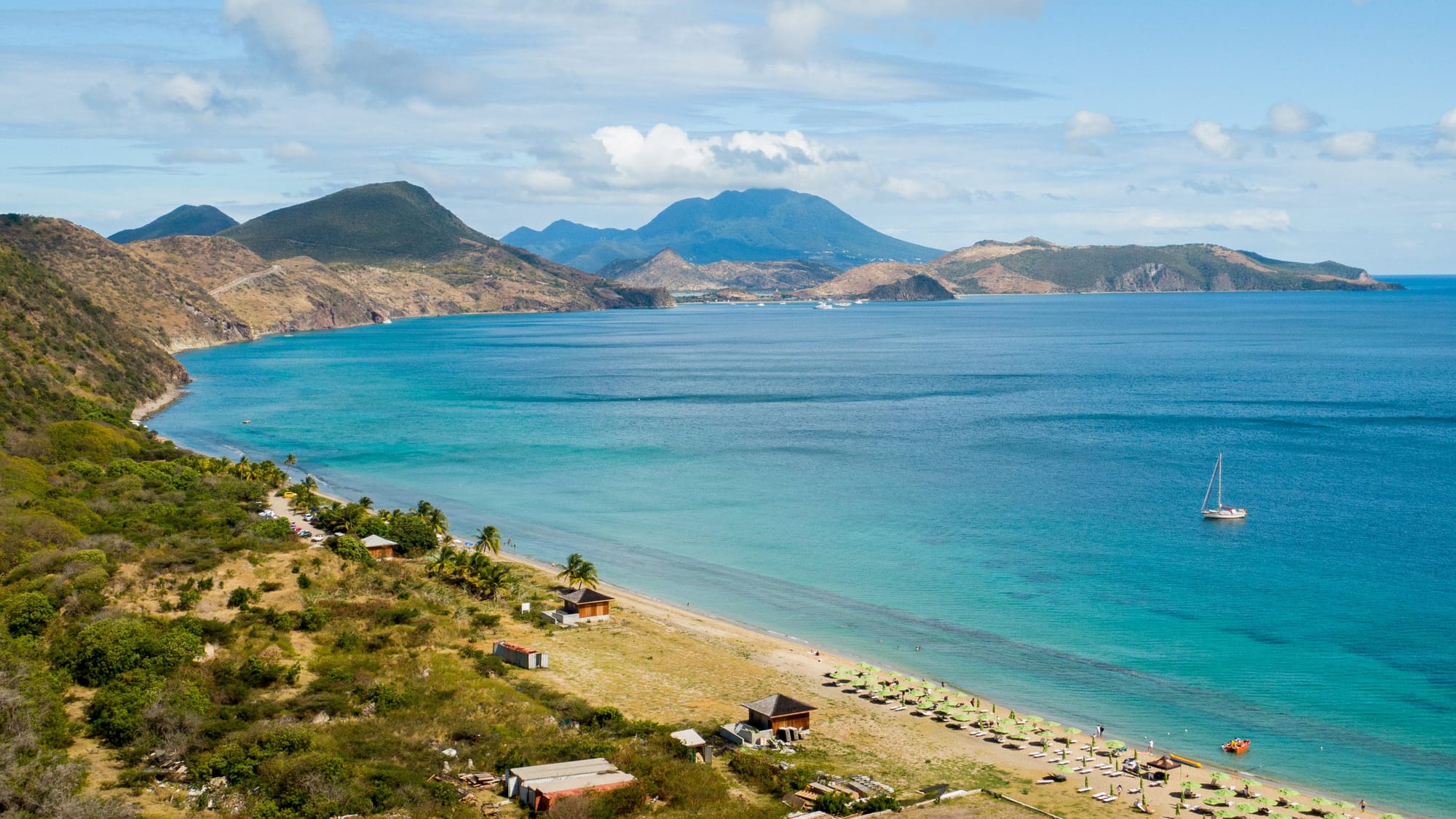View of South Friars Bay Beach with mountains in the background and a sailboat anchored near beach.