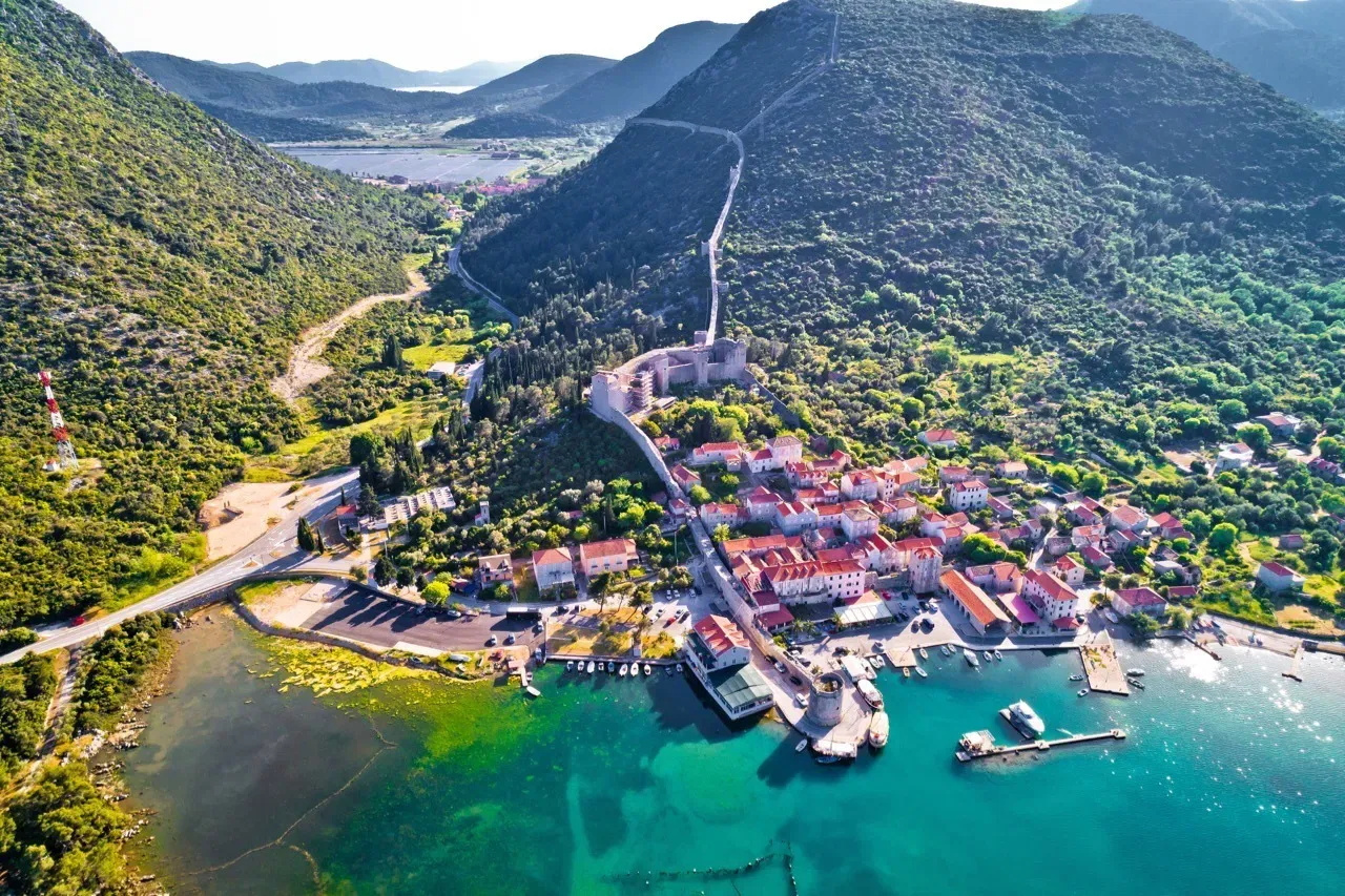 Aerial view of Ston, Croatia, showing the coastal town nestled between two green hills with the historic defensive wall running up the mountain ridge and a waterfront with docks and boats on turquoise waters.